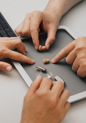 Mehrere Hände zeigen auf verschiedene Hörgeräte auf einem Tablett - Hörakustik Griesheim - Reparaturservice