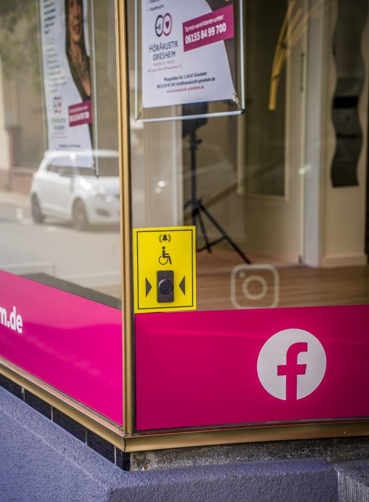 Eingang des HörAkustik Griesheim Fachgeschäfts mit barrierefreier Klingel und Werbeschildern. - örakustik Griesheim - Barrierefreier Zugang